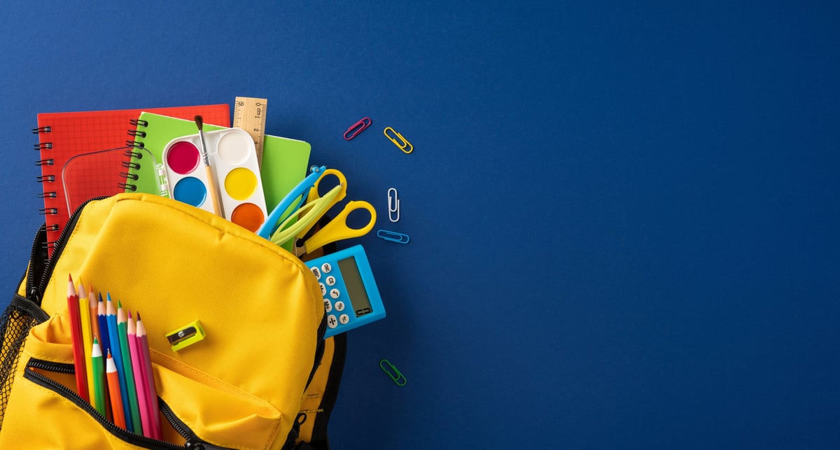 A bright yellow backpack packed with pencil, paint, scissors, calculator and notebook on a blue background evoking back to school energy and creativity