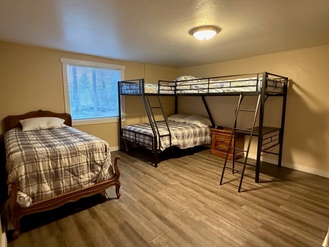 Bedroom with wooden bed on left and black metal bunk bed on right, beige walls, ceiling light, and window with natural light