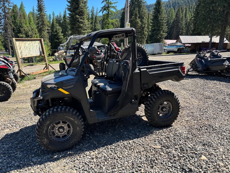 Black utility terrain vehicle parked on gravel with forested background and other ATVs visible