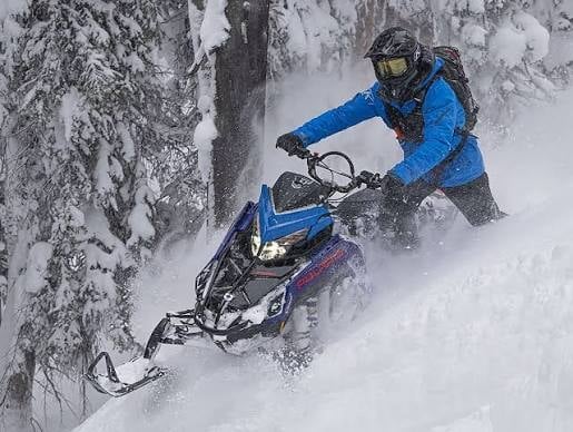 Person in blue jacket riding a blue and black snowmobile through deep snow with snow-covered trees in background