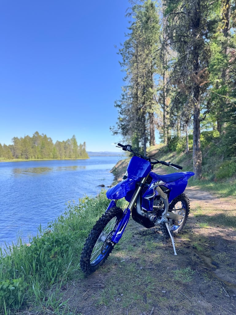 Blue dirt bike parked on a dirt trail overlooking a lake with coniferous trees and clear blue sky