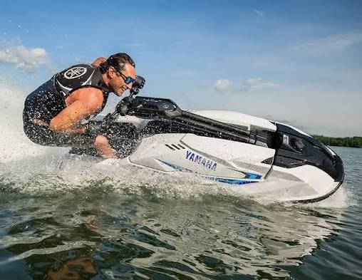 Man riding a white and black Yamaha jet ski at high speed, spraying water, on a sunny day with blue sky