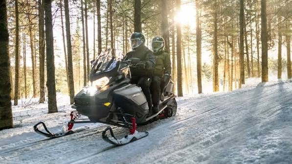 Two people riding a black and white snowmobile on a snowy forest trail lined with tall trees.