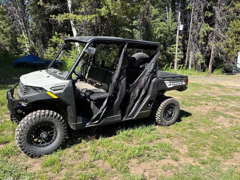 White and black side-by-side UTV with doors open parked on grass near forest trees