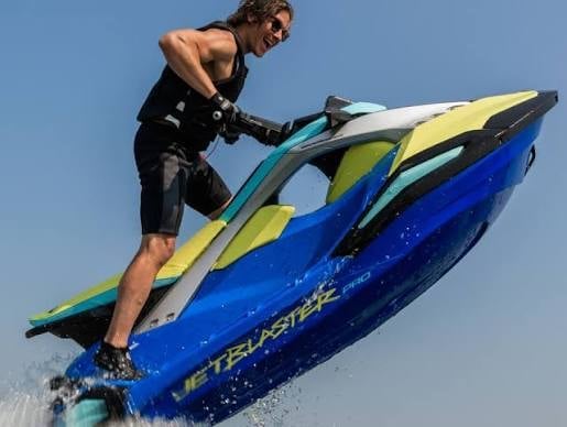 Man in wetsuit standing on a blue, yellow, and turquoise jet ski against a clear sky