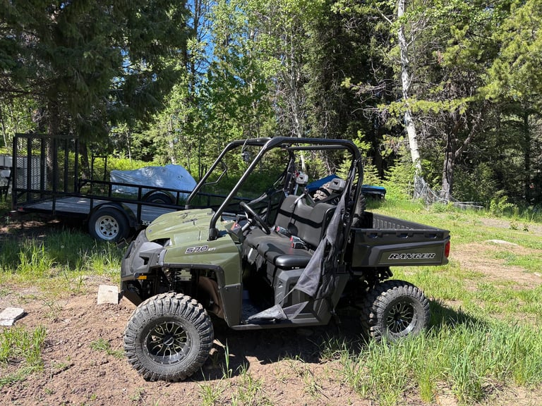 Green utility vehicle parked on dirt ground with trailer and trees in background